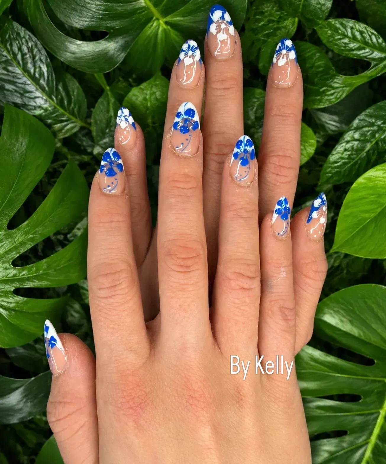 Almond nails hand-painted with blue and white hibiscus flowers on a tropical green backdrop